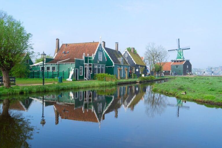 Traditional green wooden houses line a calm canal at Zaanse Schans in the Netherlands. A windmill stands in the distance and the still water reflects the homes and blue sky.