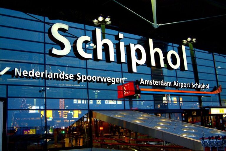 Glass facade of Schiphol Airport with large white "Schiphol" lettering above the entrance. Signs for "Nederlandse Spoorwegen" and "Amsterdam Airport Schiphol" are visible on the building, with escalators and transportation facilities in the foreground.