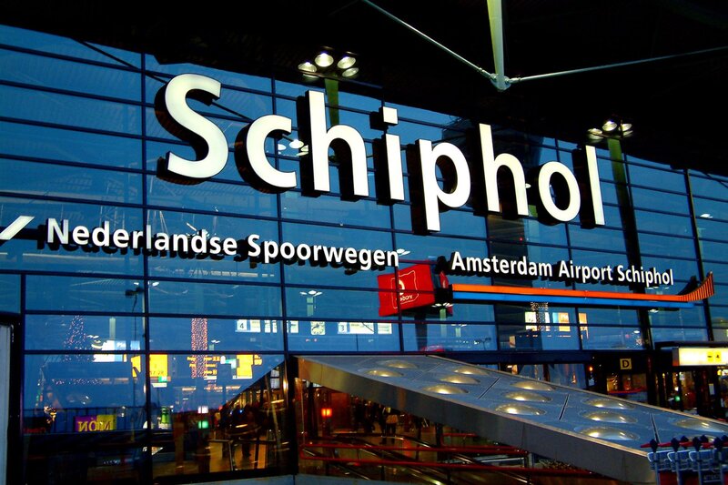 Glass facade of Schiphol Airport with large white "Schiphol" lettering above the entrance. Signs for "Nederlandse Spoorwegen" and "Amsterdam Airport Schiphol" are visible on the building, with escalators and transportation facilities in the foreground.