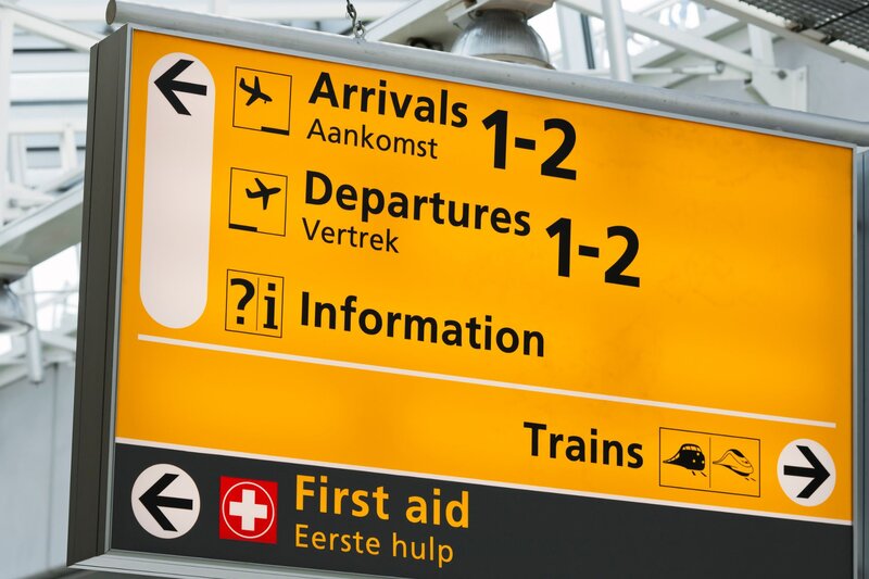 Large yellow airport wayfinding sign showing directions for "Arrivals 1-2", "Departures 1-2", "Information", "Trains", and "First aid". Black arrows and transportation icons help travelers navigate the terminal, with Dutch translations displayed beneath several headings.