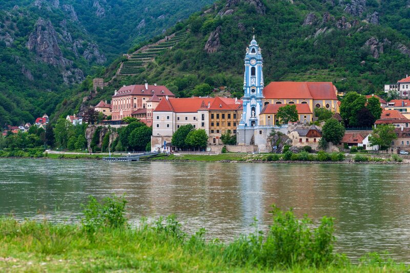Baroque monastery and church with a blue bell tower overlooking the Danube River, surrounded by steep green hills and vineyard terraces. This historic riverside setting highlights one of the best places to visit in Austria for scenic landscapes and cultural landmarks.