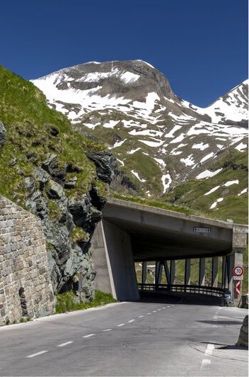 Mountain road passing through a protective tunnel beneath rugged alpine peaks streaked with late season snow. The dramatic high altitude scenery reflects the spectacular drives and landscapes found in Austria.