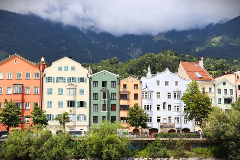 Row of pastel coloured historic buildings lining a riverbank with forested mountains rising behind them under low clouds. The charming architecture and alpine backdrop showcase the character of Austrian mountain towns.