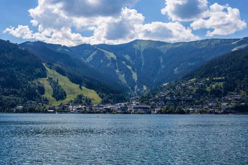 Wide view across a clear lake toward a village nestled between green mountain slopes under a sky filled with white clouds. The combination of water, mountains, and traditional settlements makes this one of the best places to visit in Austria for outdoor scenery.