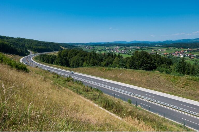 Modern highway curving through rolling farmland, forests, and small villages in the Slovenian countryside under a clear blue sky. The well maintained road network makes driving in Slovenia an easy way to explore the country's rural regions.