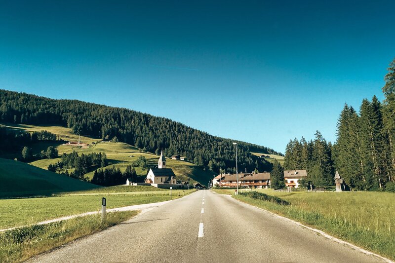 Two lane road leading toward a small Slovenian village with a church steeple, surrounded by meadows and forested hills. This peaceful countryside route reflects the scenic drives and charming communities found throughout Slovenia.
