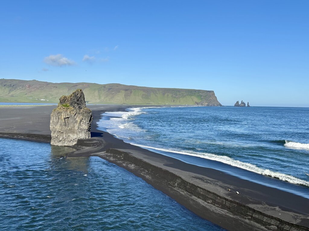 Black sand beach and dramatic sea stacks along Iceland's southern coast with waves rolling onto the shoreline under a bright blue sky. This wide coastal landscape showcases one of the most memorable sights to visit in Iceland with kids.