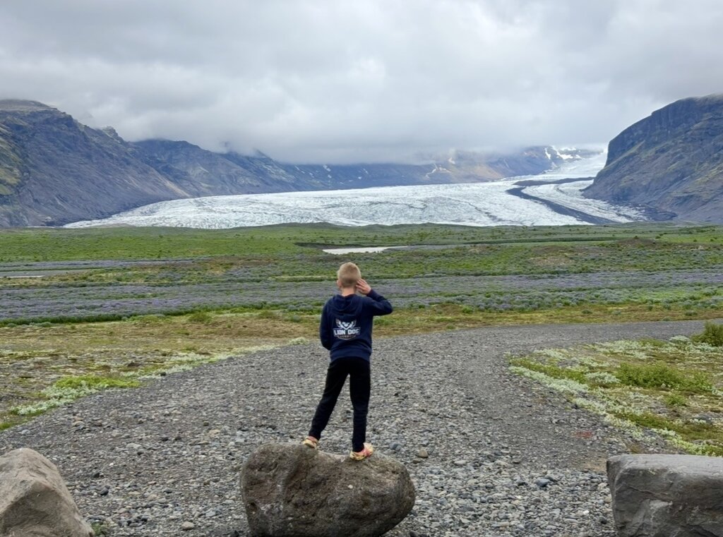 Child standing on a large rock overlooks a glacier stretching through a mountain valley in Iceland beneath low clouds. The expansive view demonstrates the awe inspiring natural landscapes families can experience in Iceland with kids.