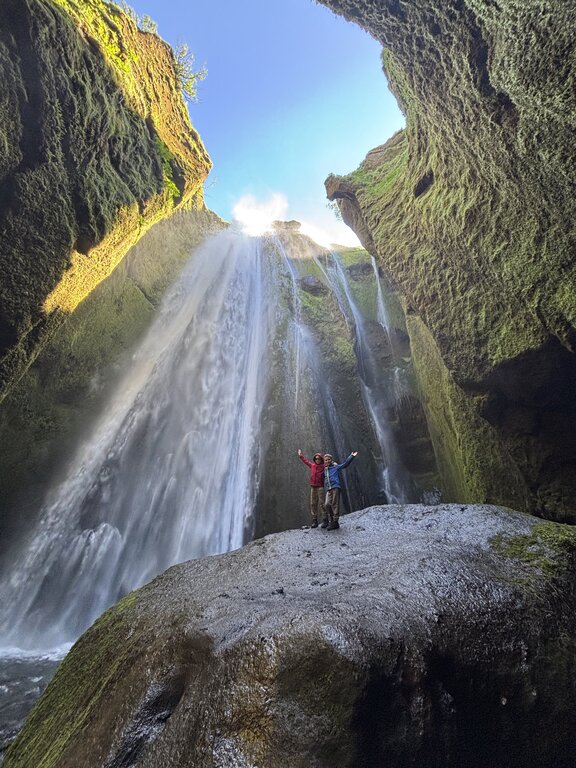 Two children stand on a large rock beneath a towering waterfall surrounded by moss covered canyon walls in Iceland. Sunlight streams through the opening above, highlighting the scale and adventure of exploring Iceland with kids.