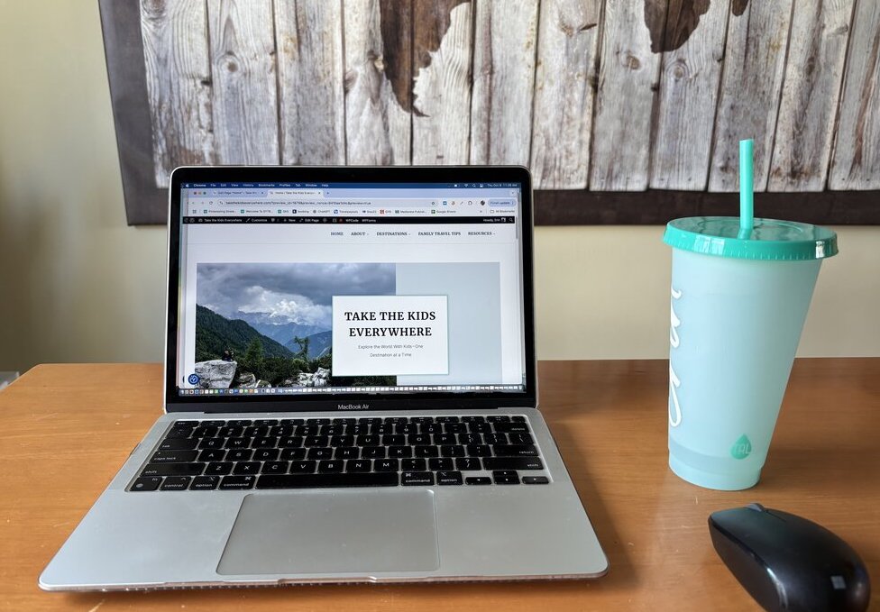 Open laptop displaying the "Take the Kids Everywhere" travel blog homepage on a dining room table beside a teal reusable cup and computer mouse. The photo shows the everyday workspace behind running a travel blog, helping illustrate whether travel blogging is worth it.