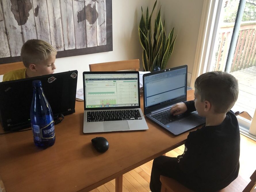 Two children sit at a dining room table using laptops for online work while a third laptop sits open between them. The scene shows a family balancing remote work and learning from home, illustrating that travel blogging can be managed from anywhere.