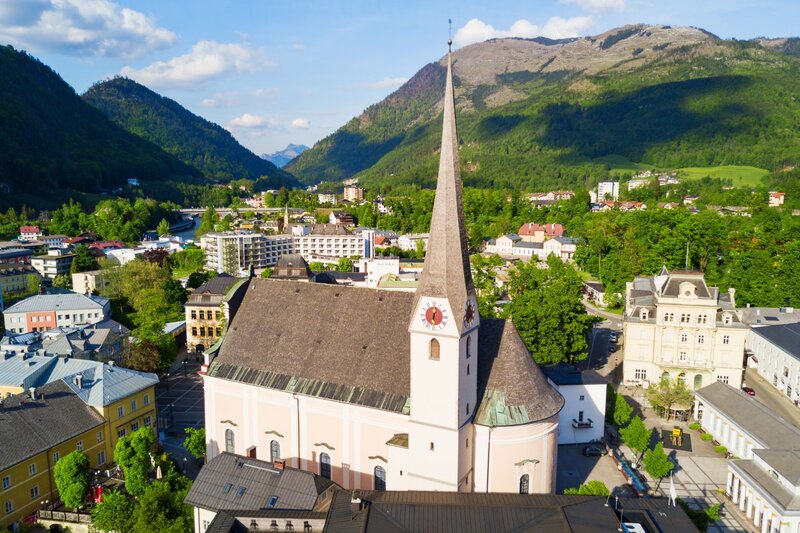 Aerial view of the parish church in Bad Ischl with its tall steeple rising above the town center, surrounded by green alpine hills and mountain peaks. The historic spa town is one of the best places to visit in Austria, known for its imperial heritage and scenic Salzkammergut setting.