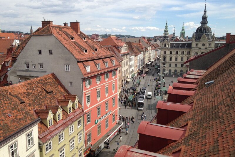 View over the red tiled rooftops of Graz's Old Town with a tram traveling along a busy pedestrian street lined with historic buildings. The city's mix of Renaissance, Baroque, and modern architecture makes Graz one of the best places to visit in Austria for culture, history, and urban sightseeing.