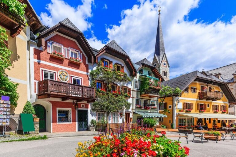 Colourful traditional houses decorated with flower boxes line a square in Hallstatt, with the town's church steeple rising above the rooftops against a blue sky with scattered clouds. This picturesque lakeside village in the Salzkammergut region is one of the most popular places to visit in Austria for its historic charm and alpine scenery.