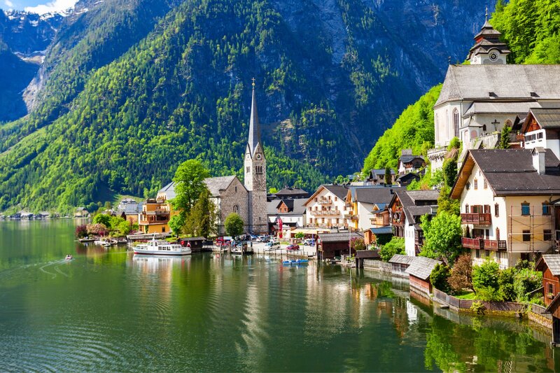 Lakeside village with a tall church steeple, traditional alpine houses, and steep forested mountains reflected in calm water. This picturesque setting is one of Austria's most recognizable destinations and a popular stop for travellers.