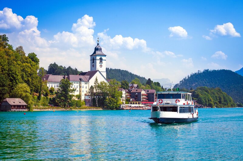White passenger boat crossing a bright blue lake with a historic church and lakeside buildings visible in the background. The peaceful waterfront scene captures the beauty of Austria's lake regions and cultural heritage.