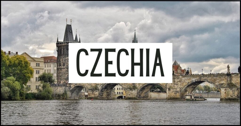 Prague's Charles Bridge spans the Vltava River beneath a dramatic cloudy sky, with Gothic towers and historic buildings lining the waterfront. Large white text overlay reads "CZECHIA".