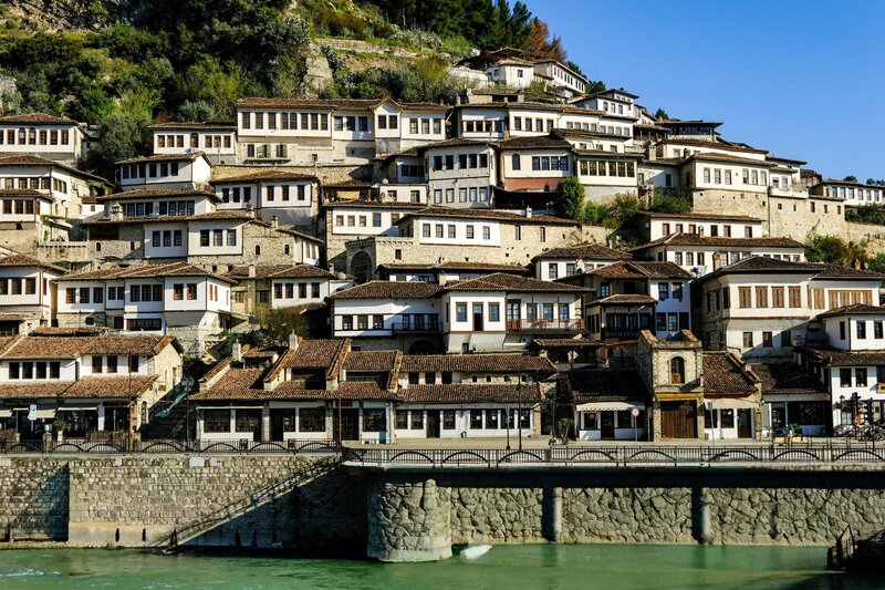 Traditional white Ottoman-era houses cascade down the hillside in Berat, Albania, overlooking the river and stone embankment below. The tightly clustered homes with dark tiled roofs showcase the city's distinctive architecture, making Berat one of Europe's most picturesque hidden gems.