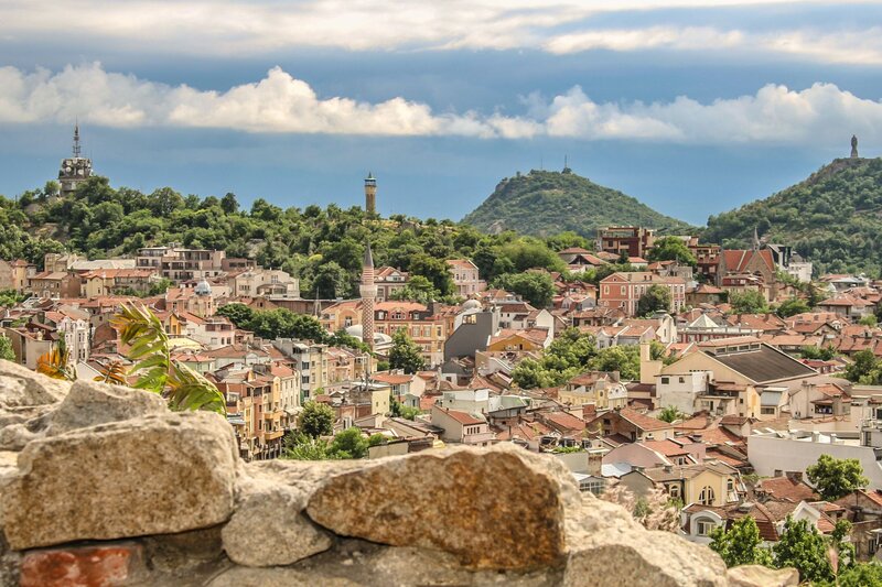 Panoramic view of Plovdiv with red tiled rooftops, a mosque minaret, and rolling green hills surrounding the historic city. Stone walls frame the foreground while dramatic clouds stretch across the sky above the skyline.