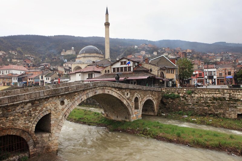 Historic stone bridge spanning a fast flowing river in Prizren, Kosovo with a mosque featuring a large dome and tall minaret rising behind it. Traditional buildings line the riverbank, with wooded hills and scattered homes forming the backdrop beneath an overcast sky.