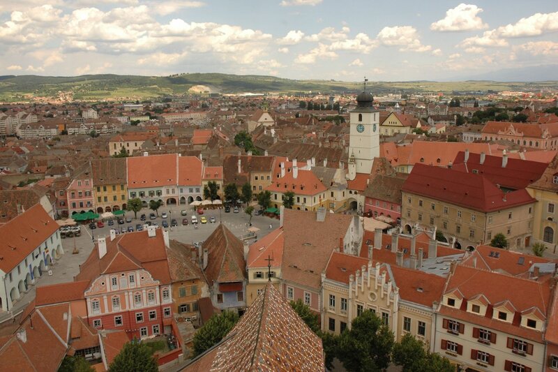 : Elevated view of Sibiu, Romania, featuring its red tile rooftops, colorful historic buildings, and the iconic clock tower overlooking the main square. Rolling green hills and a sky filled with scattered clouds frame this hidden gem in Europe, highlighting the city's well-preserved medieval architecture.
