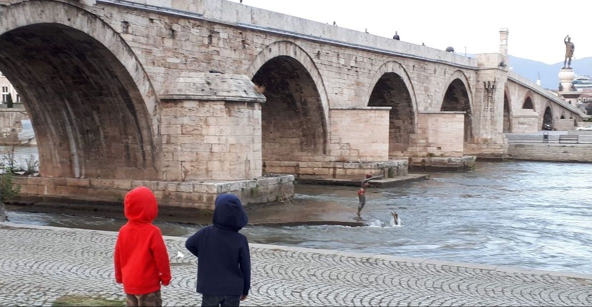 wo children in red and navy hooded sweatshirts stand on the riverbank looking at a statue of a person jumping into the Vardar River beside the historic Stone Bridge in Skopje. The bridge's large stone arches stretch across the water, with statues and city landmarks visible in the background beneath an overcast sky.