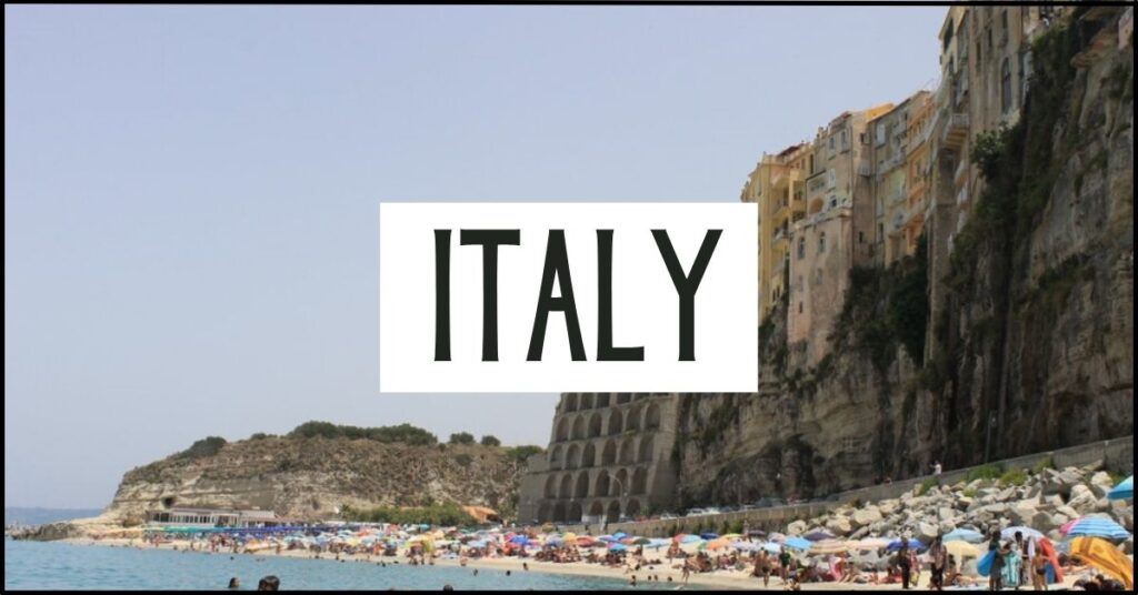 Beach lined with colourful umbrellas beneath dramatic cliffside buildings overlooking the sea in Calabria Italy. Large white text overlay reads "ITALY".