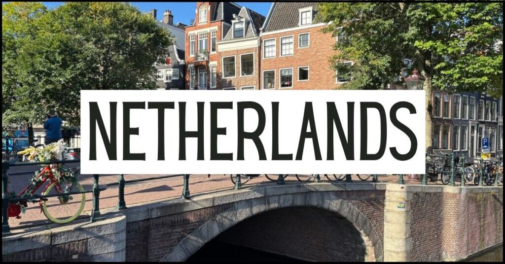 Brick canal bridge lined with bicycles crossing one of Amsterdam's tree lined canals, with traditional narrow houses in the background. Large white text overlay reads "NETHERLANDS".