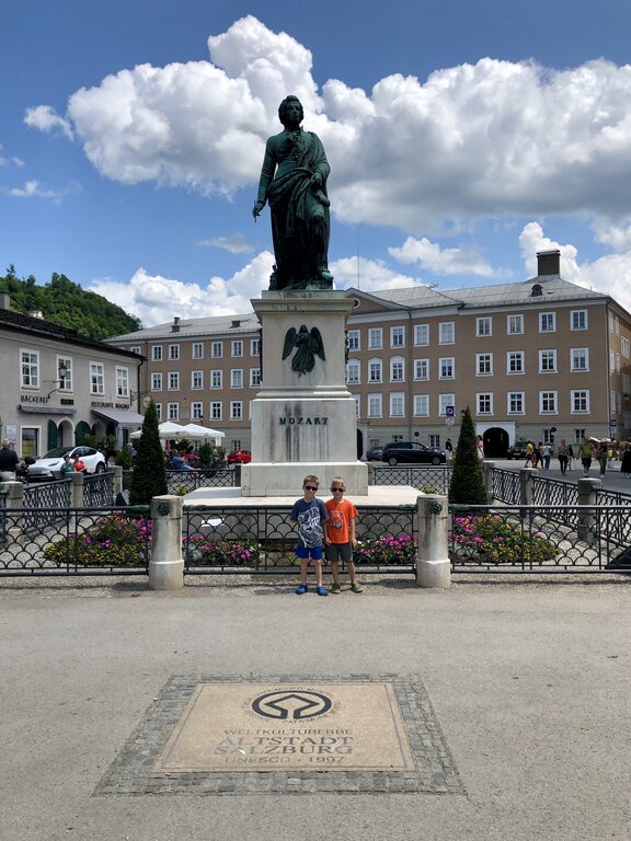 Two children stand in front of the Mozart monument in Mozartplatz with flower beds and historic buildings behind them. A UNESCO World Heritage marker is visible in the foreground, making this a memorable stop when exploring Salzburg with kids.