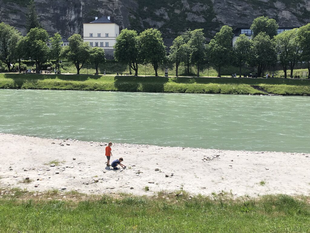 Two children play with stones on the sandy bank of the Salzach River while green trees line the opposite shore.