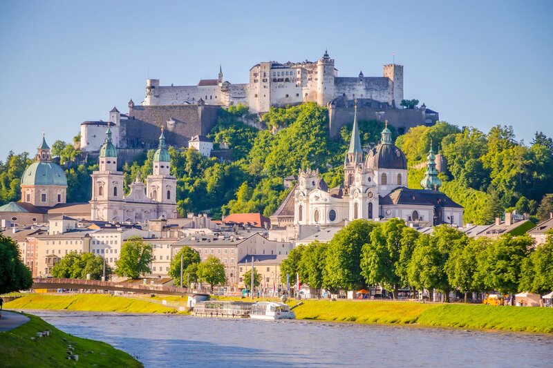 Panoramic view of Salzburg with the Salzach River in the foreground, cathedral domes across the skyline, and Hohensalzburg Fortress overlooking the city from the hilltop. This iconic view showcases one of the highlights of Salzburg with kids.