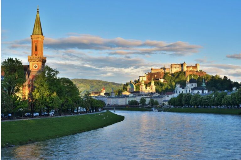 Evening view along the Salzach River with historic church towers and Hohensalzburg Fortress illuminated by warm golden light. The scenic riverside is a beautiful place to end a day exploring Salzburg with kids.