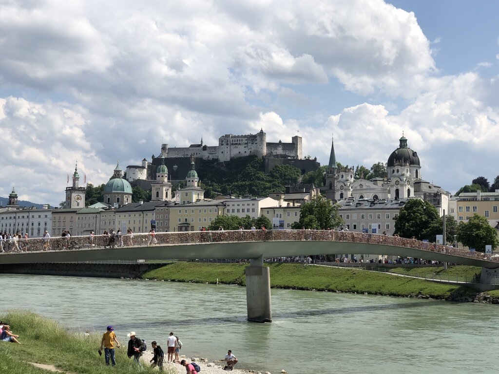 View across the Salzach River toward the pedestrian bridge and Hohensalzburg Fortress towering above the city skyline. People relax along the riverbank while others cross the bridge.