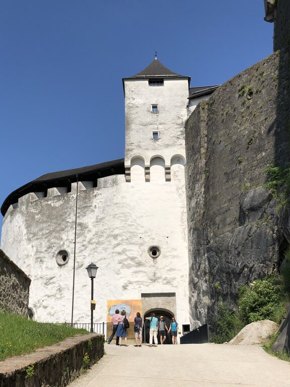 Visitors walk toward the white stone entrance of Hohensalzburg Fortress beneath tall defensive walls and a square tower. The uphill approach gives a sense of the fortress's impressive scale.