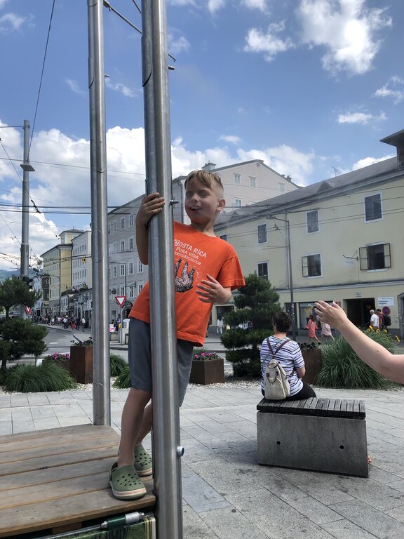 Young boy in an orange T shirt smiles while climbing on a metal structure with mist spraying from the side in a public square in Salzburg, surrounded by historic buildings.