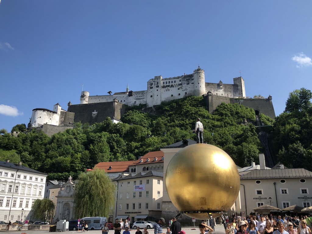 Hohensalzburg Fortress rises above Salzburg while people gather around the large golden Sphaera sculpture in Kapitelplatz.