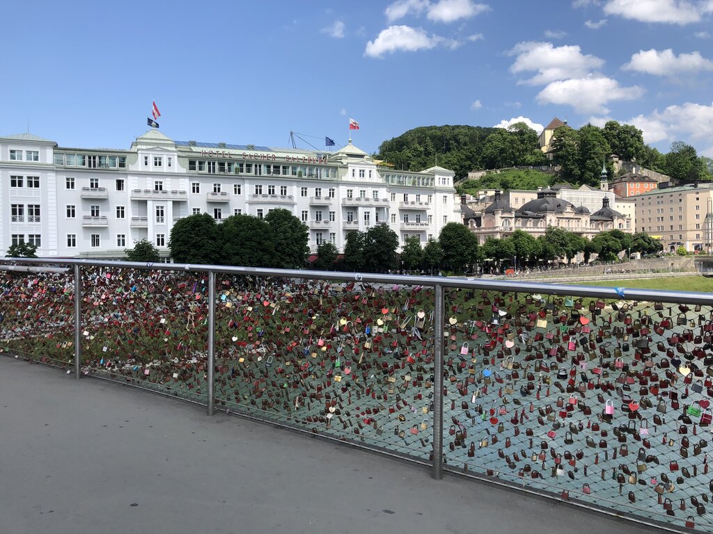 Bridge covered with hundreds of colorful love locks stretches across the Salzach River with grand historic buildings in the background. The scene captures a popular riverside walk through Salzburg.
