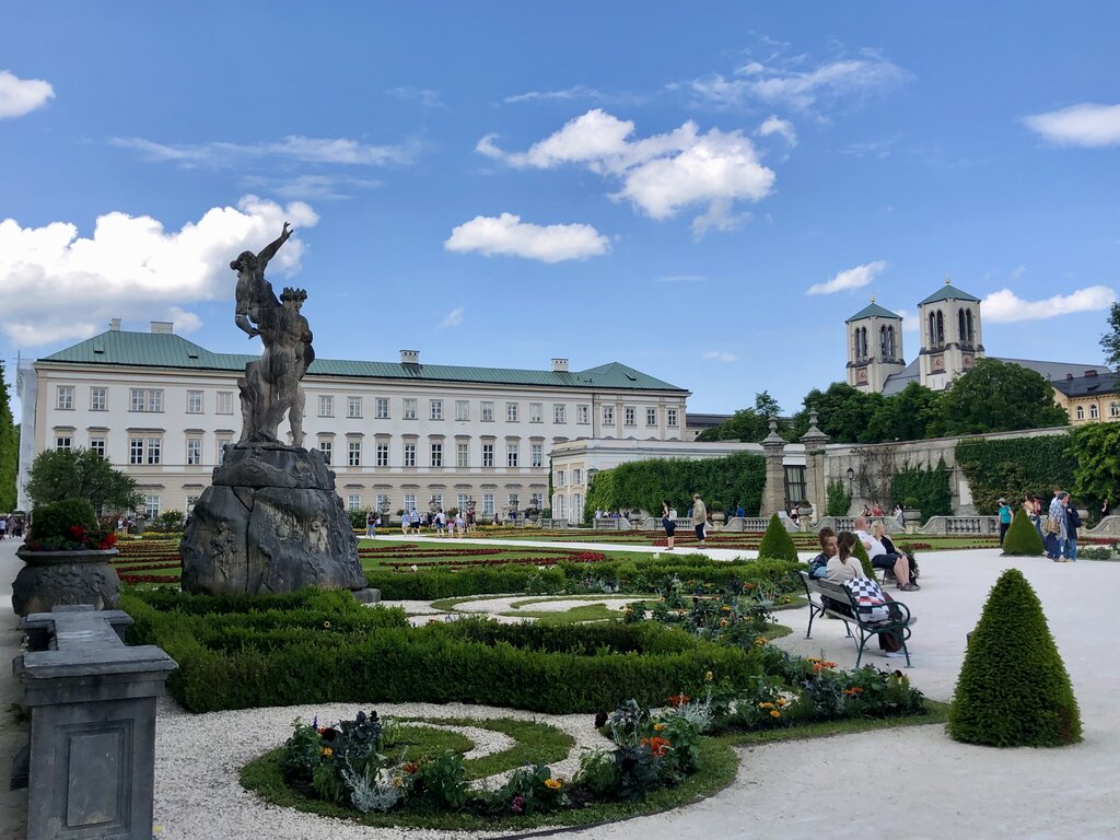 Formal gardens at Mirabell palace in Salzburg, Austria, with colourful flower beds, trimmed hedges, and a large statue surrounded by visitors. Church towers and historic buildings rise beyond the landscaped grounds.