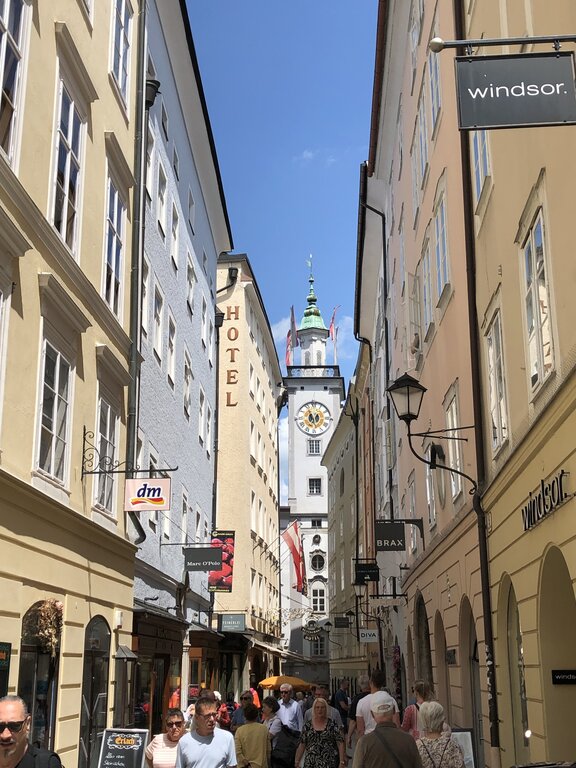 Busy shopping street lined with historic pastel buildings leading toward the clock tower of Salzburg's Old Town. Shops, cafés, and pedestrians fill the narrow street on a sunny day.