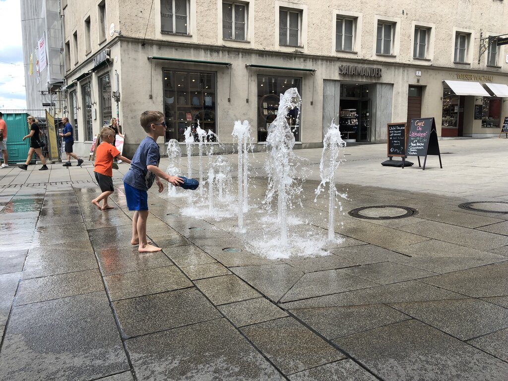 Two children play barefoot in a splash fountain on a pedestrian street while water jets spray into the air around them. Cooling off at interactive fountains is a fun way to enjoy Salzburg with kids on a warm day.