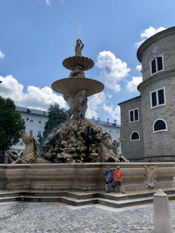 Two children pose beside the ornate Residenz Fountain with sculpted horses, stone figures, and a tiered fountain beneath a bright blue sky. The impressive fountain is one of Salzburg's most recognizable landmarks.