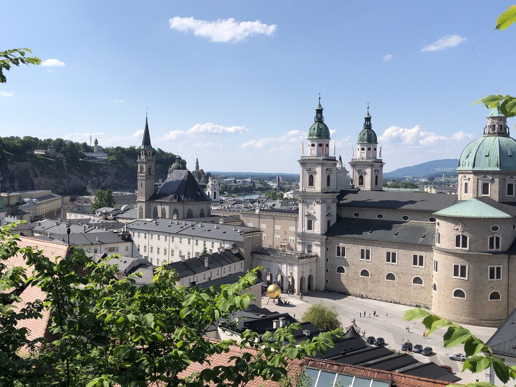 Panoramic view across Salzburg featuring cathedral domes, church spires, and historic rooftops beneath a bright blue sky. Green leaves frame the foreground from a hillside viewpoint.