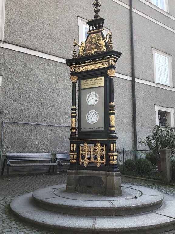 Ornate black and gold historic weather pillar standing on a circular stone base beside a grey building. The display features two dials and the text "Stadtverein Salzburg" and "Stadttheater Salzburg."