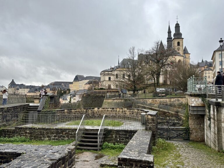 View of Luxembourg City's historic center with stone fortifications, bridges, and church spires under a dramatic overcast sky. Layered historic buildings and tree lined terraces overlook the lower valley, highlighting the city's unique landscape and architecture.