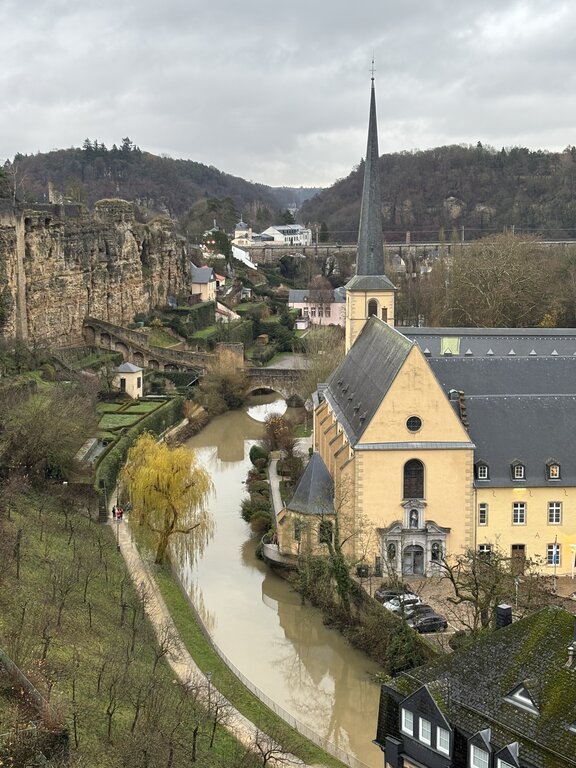 Elevated view of Luxembourg City's Grund district with a pale stone church, winding river, arched bridges, and dramatic sandstone cliffs surrounded by wooded hills. Historic buildings and walking paths follow the river beneath an overcast sky, showcasing the city's distinctive natural setting and architecture.