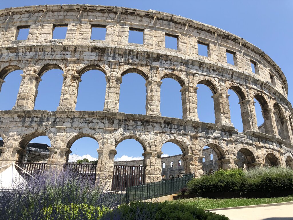 Close up view of the Pula Arena showing weathered limestone arches and multiple tiers of the ancient Roman amphitheater against a clear blue sky. Lavender plants and green shrubs line the foreground, emphasizing the scale and preserved stone architecture.