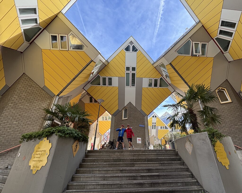 Bright yellow Cube Houses in Rotterdam, Netherlands, viewed from below with their tilted architecture forming a geometric frame around a blue sky. Two boys stand at the top of a wide staircase beneath the houses, while palm trees, bicycles, and surrounding walkways highlight the distinctive urban design and scale of the space.