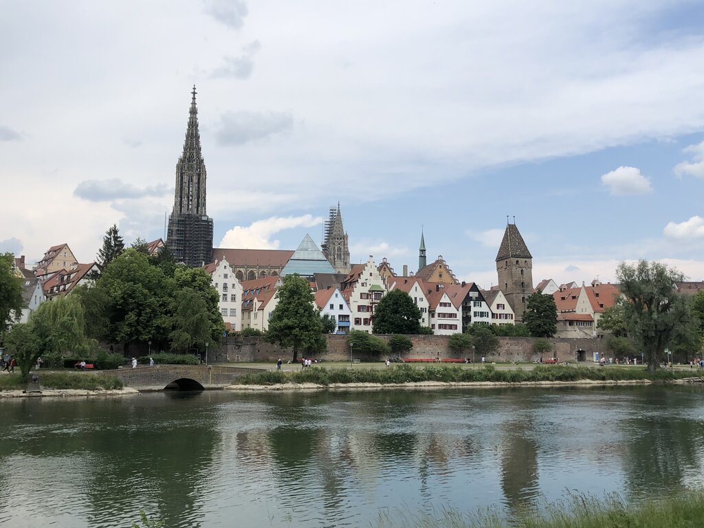 Skyline of Ulm viewed from across the Danube River with the towering Gothic spire of Ulm Minster rising above colorful historic buildings and mature trees. Calm water in the foreground reflects the cityscape beneath a cloudy sky with patches of blue.