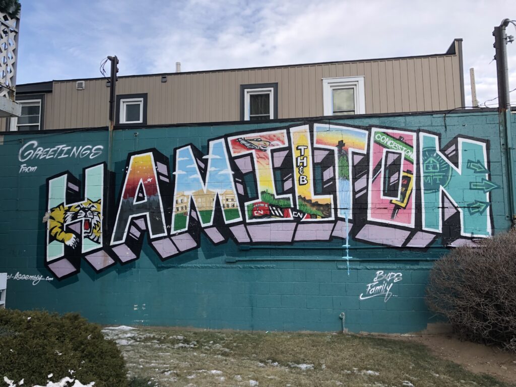 Colourful mural on a building wall with the word "HAMILTON" filled with local landmarks and scenes. The mural also includes the painted text "Greetings from," "TH & B," "Concession," and "BuffB Family."