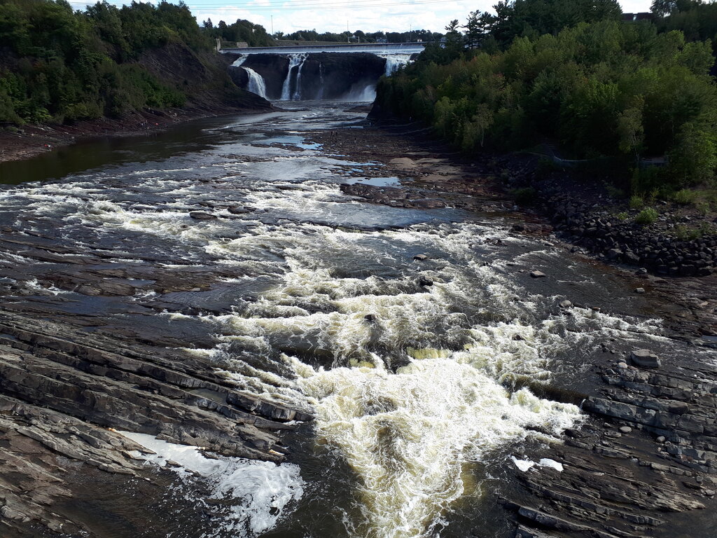 Whitewater rapids rush over layered dark rocks toward a broad waterfall surrounded by dense green forest. The view shows the rocky river and powerful flowing water at Chutes de Chaudiere near Quebec City.